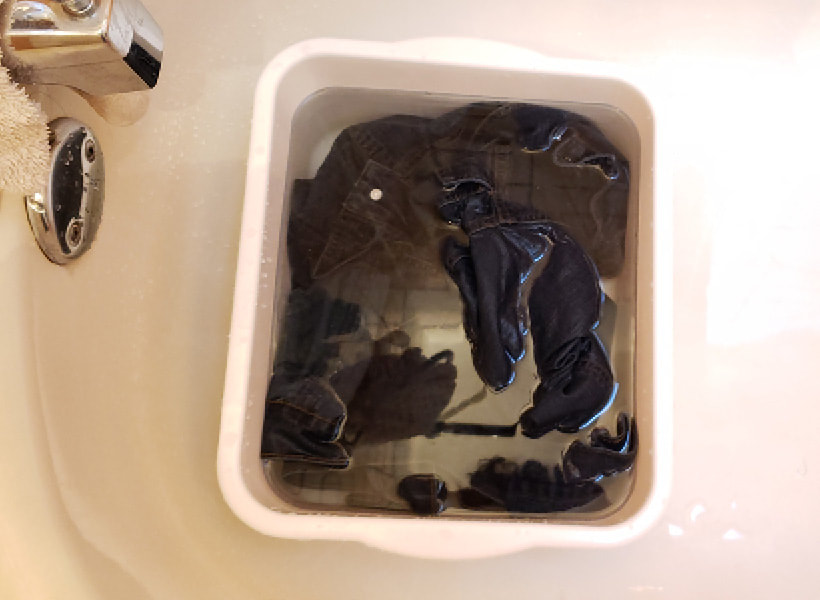 The dark jacket is submerged in water and bleach in the white bucket. The bucket is inside a bathtub.