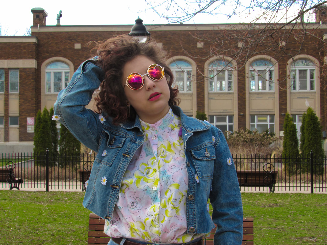Shot of the model from the waist up in front of a brick building and park benches. She is facing the camera with the jacket unbuttoned. She is wearing pink and yellow sunglasses with a neutral expression. Her hand is running through her hair.
