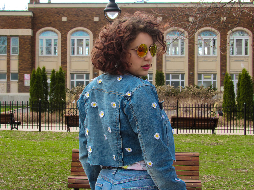 Shot of the model from the waist up in front of a brick building and park benches. She is turned slightly to the right to expose the jacket's back and sleeves. She is wearing pink and yellow sunglasses and pouting with her chin down.