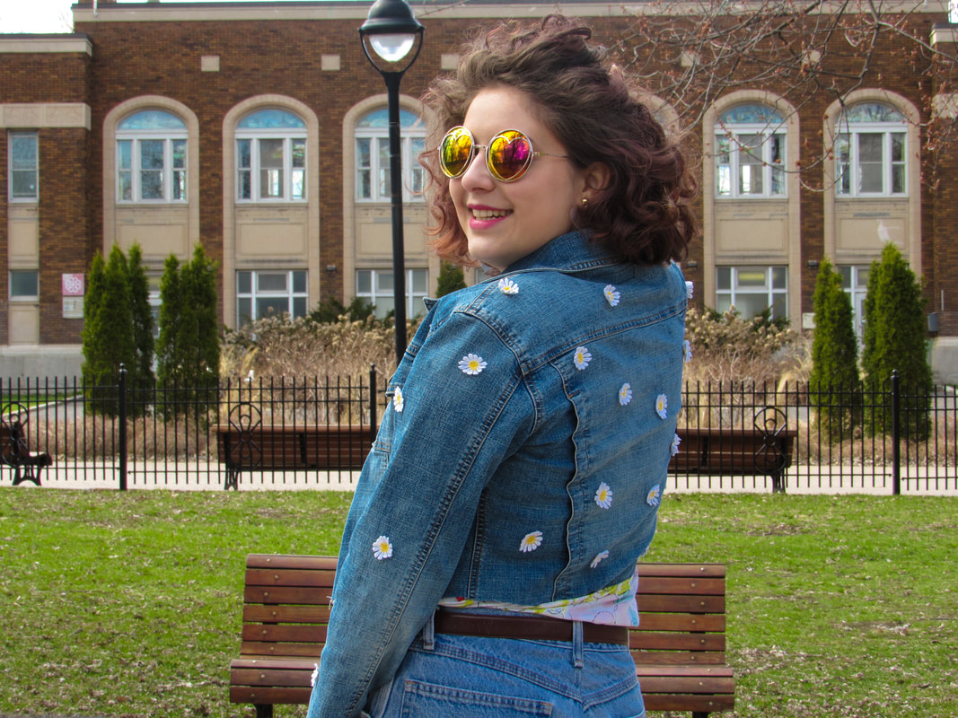 Shot of the model from the waist up in front of a brick building and park benches. She is turned slightly to the left to expose the jacket's back and sleeves. She is wearing pink and yellow sunglasses and smiling.