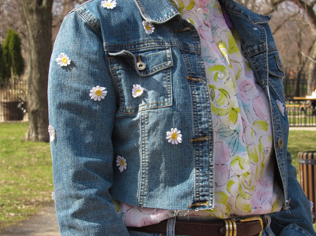 Closeup of denim jacket covered in daisy appliqués. Model is wearing it over a pink and yellow floral button up. She's in the park and her face is out of frame.