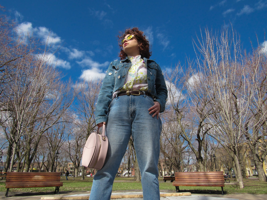 Full body shot of model in the park. She is wearing the jacket, the button-up shirt, the sunglasses, light-wash jeans, and a round pink purse. She is looking off to the side.